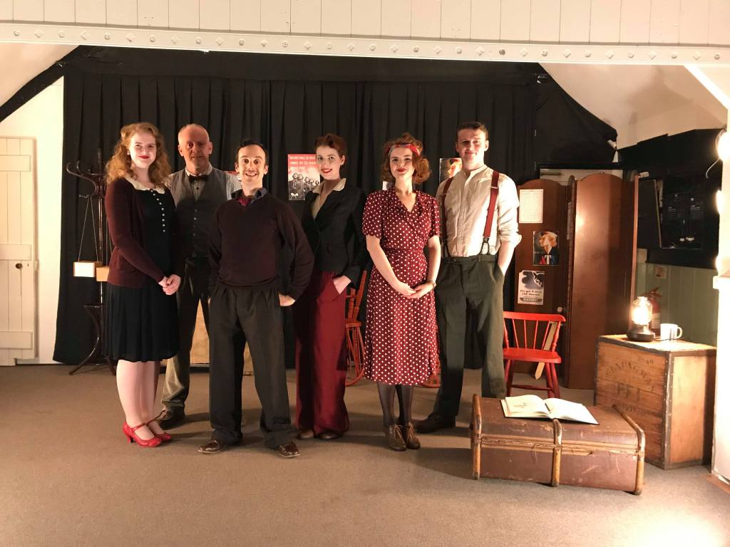 The cast of Greyhounds in 1940s costumes standing on stage in a village hall.