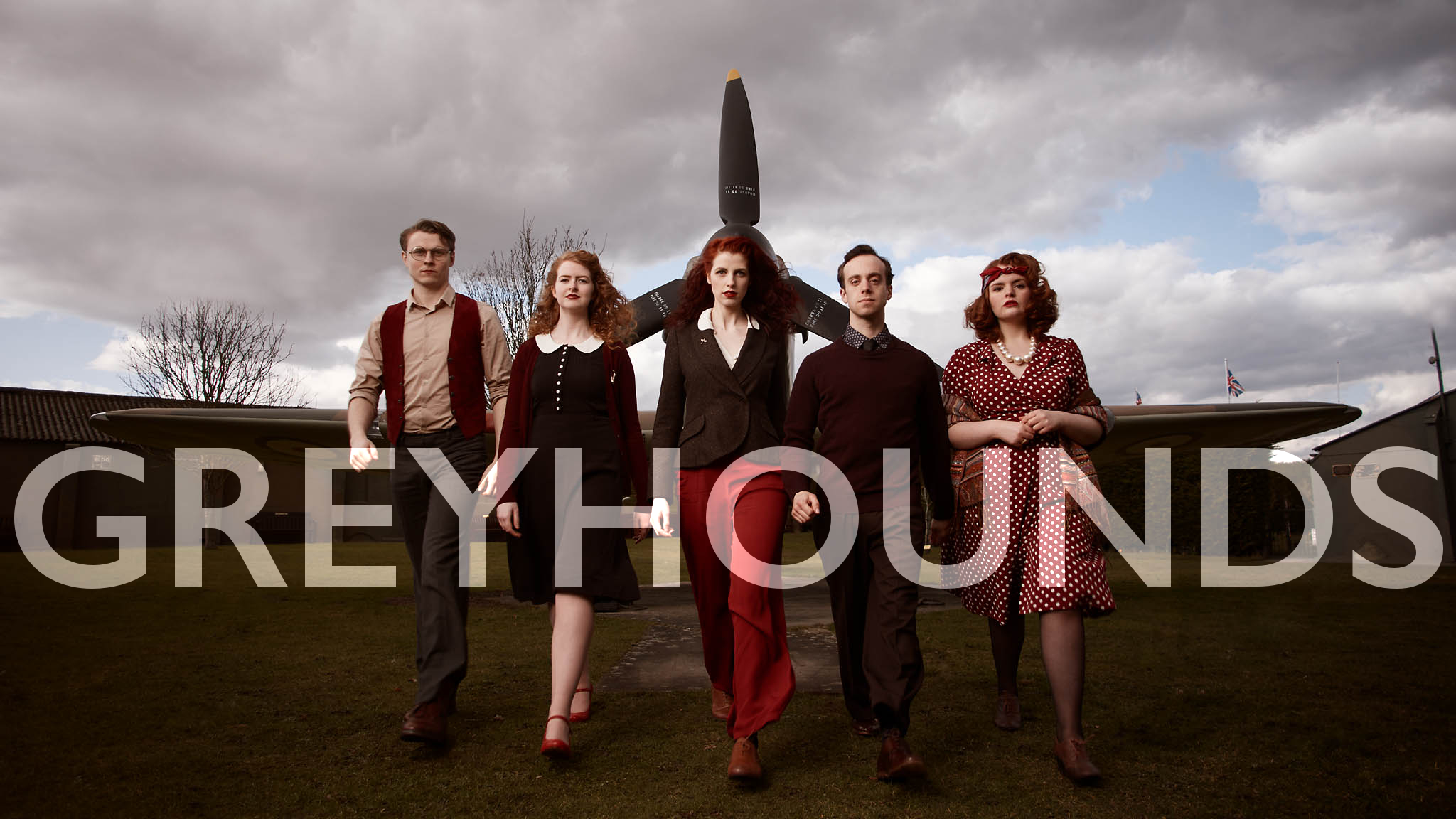 The cast of Greyhounds, wearing 1940s costumes, striding out in front of a World War Two Hurricane, overlaid with the words 'GREYHOUNDS'.
