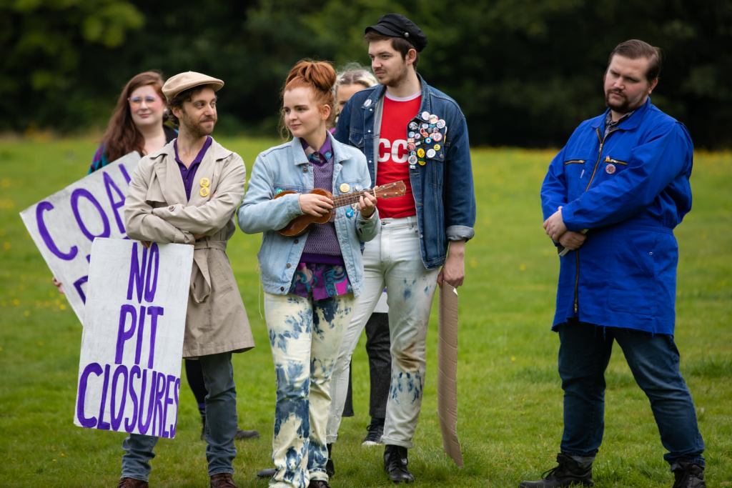 The Mechanicals from Time & Again's production of A Midsummer night's Dream. They are holding large signs saying 'No Pit Closures' and 'Coal not Dole' and dressed mainly in denim.