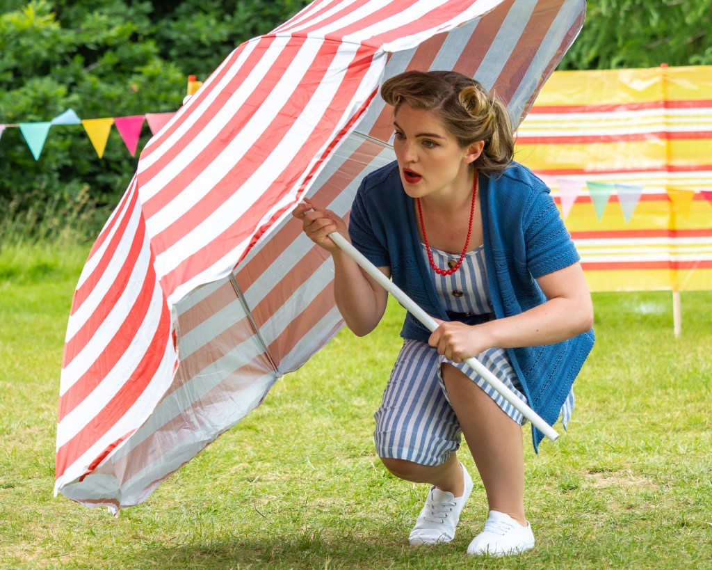 Beatrice in Time & Again's production of Much Ado About Nothing. She is hiding behind a red striped umbrella and wearing a blue 1950s playsuit.