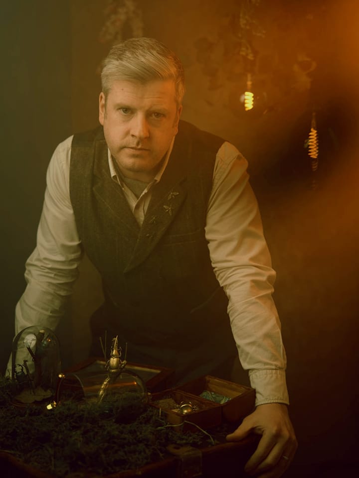 Ben Hynes in Earwig, wearing a tweed waistcoat, cream shirt, and green cravat. He's leaning on a bench of entomology equipment and looking serious.